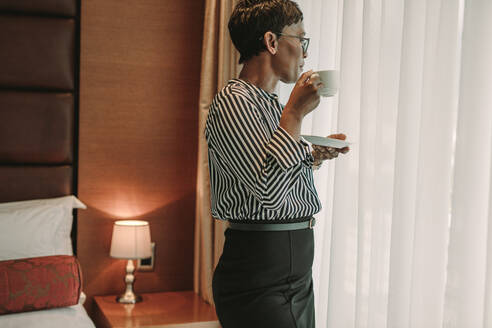Businesswoman standing near in hotel room window with a cup of coffee. Mature woman in formals drinking coffee in hotel room. - JLPSF16131