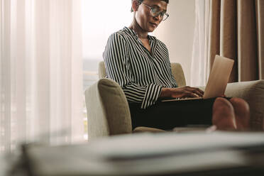 African woman in formal clothes sitting on armchair in hotel room using laptop. Businesswoman on business trip working from hotel room. - JLPSF16136