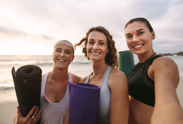 Nahaufnahme von glücklichen Frauen am Strand, die zusammen stehen und Yogamatten halten. Drei Yoga-Frauen stehen am Strand. - JLPSF16150