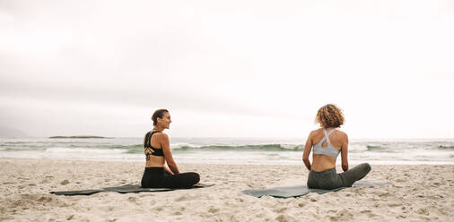 Rear view of two women in fitness wear practicing yoga sitting on the beach. Fitness women sitting on yoga mats doing yoga facing the sea. - JLPSF16159