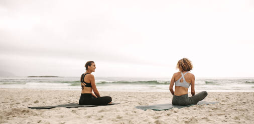 Rückansicht von zwei Frauen in Fitnesskleidung, die am Strand sitzend Yoga üben. Fitness-Frauen sitzen auf Yogamatten und machen Yoga mit Blick auf das Meer. - JLPSF16159