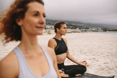 Frauen in Fitnesskleidung machen Yoga am Strand sitzend an einem bewölkten Tag. Frau sitzend in Yoga-Position mit den Händen auf den Knien ruhend. - JLPSF16161