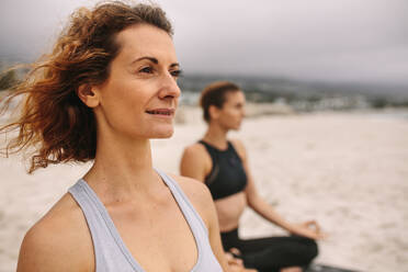 Nahaufnahme einer Frau, die zusammen mit ihrer Freundin am Strand Yoga übt. Frauen in Fitnesskleidung am Strand, die an einem bewölkten Tag Yoga machen. - JLPSF16162