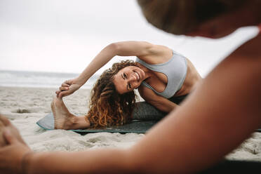 Fitness-Frauen sitzen auf Yogamatten am Strand und machen Stretching-Workout. Frauen üben Dehnungshaltungen des Yoga sitzen in der Nähe des Meeres. - JLPSF16163