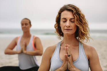 Zwei Frauen sitzen am Strand und meditieren mit geschlossenen Augen und zusammengelegten Händen. Fitness-Frauen machen Yoga am Strand an einem bewölkten Morgen. - JLPSF16165