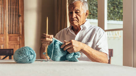 Senior man knitting warm clothes with wool yarn on table. Retired man making a sweater with needles and wool yarn at home. - JLPSF16168