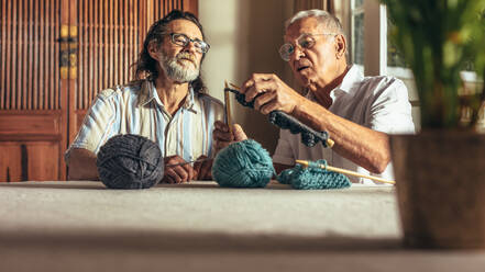 Älterer Mann, der seinen Freunden das Stricken von Wollkleidung beibringt. Älterer Mann, der von seinen Freunden, die zu Hause sitzen, das Stricken von Wollkleidung lernt. - JLPSF16172