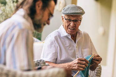 Senior man with hat looking at his friend doing knitting while sitting at patio. Elderly male friends kitting together. - JLPSF16190