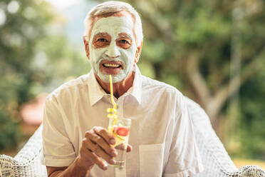 Lustiger älterer Mann mit Tonmaske im Gesicht, der Saft trinkt. Älterer Mann im Ruhestand, der zu Hause seine Haut im Alter pflegt. - JLPSF16207