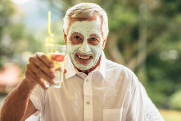 Lustiger älterer Mann mit einer Gesichtsmaske aus Ton und einem Glas Saft in der Hand, der sich im Ruhestand einer Spa-Gesichtsbehandlung unterzieht. - JLPSF16208