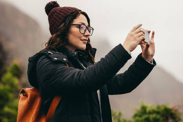 Frau in warmer Kleidung, die mit ihrem Smartphone auf einem Berggipfel steht und die Landschaft mit ihrem Mobiltelefon aufnimmt. - JLPSF16227