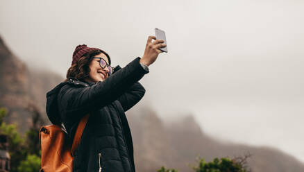 Glückliche junge Frau im Winterurlaub, die ein Selfie macht. Sie hält das Handy hoch und posiert, während sie auf einem Berggipfel steht. - JLPSF16228
