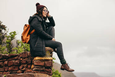 Junge Frau, die auf einer Steinmauer am Berghang sitzt und die Aussicht betrachtet. Weibliche Touristin, die sich auf einem Berg ausruht und die Aussicht betrachtet. - JLPSF16230