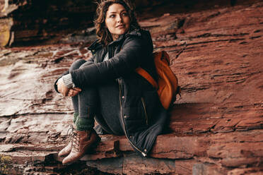 Frau in warmer Jacke, die auf einem felsigen Berg sitzt und wegschaut. Kaukasische weibliche Touristin, die die Aussicht vom Berg betrachtet. - JLPSF16235