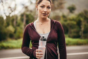 Fitness-Frau hält eine Wasserflasche und schaut in die Kamera. Läuferin entspannt sich nach dem Training am Morgen. - JLPSF16243