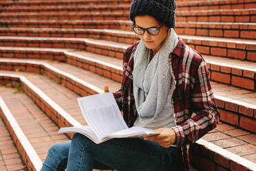 Schöne Studentin mit Lehrbüchern auf dem College-Campus. Junge kaukasische Frau mit Brille sitzt auf den Stufen und liest ein Buch. - JLPSF16249