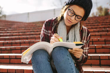 Eine Studentin benutzt einen Stift, um Text in einem Buch zu markieren. Eine Studentin sitzt auf einer Treppe auf dem Campus und studiert. - JLPSF16250