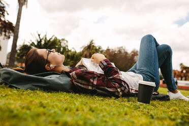 Studentin mit Kopfhörern, die mit einem Buch im Gras liegt. Mädchen ruht sich aus und hört Musik, während sie ein Buch auf dem College-Campus liest. - JLPSF16252