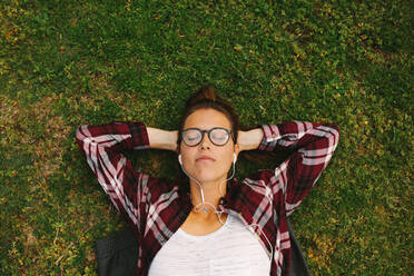 Draufsicht auf eine junge Frau mit Brille, die mit Kopfhörern Musik hört. Eine Studentin liegt mit den Händen hinter dem Kopf im Gras und entspannt sich auf dem College-Campus. - JLPSF16253