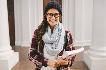 Lächelnde junge Frau mit einem Buch auf dem College-Campus. Fröhliche Studentin auf dem Universitätscampus an einem kalten Wintertag. - JLPSF16254