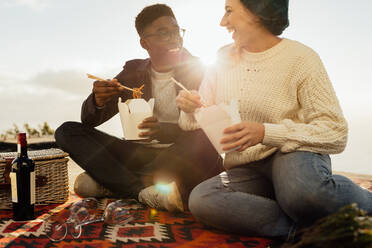 Ein gemischtrassiges Paar beim Picknick, das Nudeln isst und an einem sonnigen Tag lächelt. Ein lächelnder junger Mann und eine lächelnde Frau sitzen auf einer Picknickdecke, essen und schauen sich an. - JLPSF16264