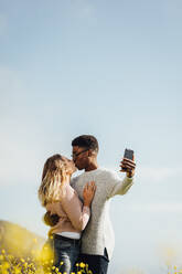 Liebevolles interrassisches Paar, das sich küsst und ein Selbstporträt im Freien macht. Romantisches Paar, das ein Selfie mit seinem Mobiltelefon macht. - JLPSF16269