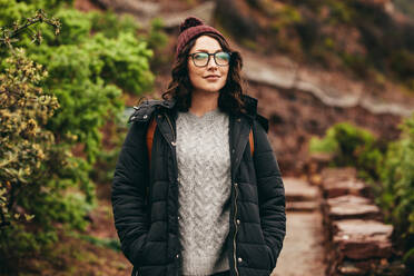 Porträt einer schönen jungen Touristin, die auf einer Steintreppe auf einem Berg steht und in die Ferne blickt. Reisende in warmer Kleidung mit Brille, die von einem Berg aus in die Landschaft blickt. - JLPSF16271