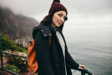 Porträt einer schönen jungen Frau, die an einem Geländer auf einem Berggipfel steht und in die Kamera schaut; eine Frau im Urlaub, die warme Kleidung trägt und eine Tasche mit sich führt. - JLPSF16272