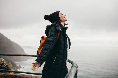 Female wearing warm jacket and hat standing by railing on mountain top with her eyes closed and arms outstretched. Woman enjoying standing in the fresh air on mountain top. - JLPSF16274