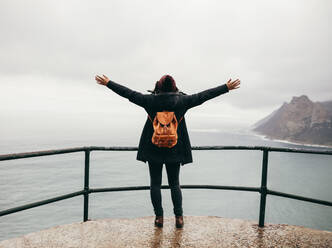 Rückansicht in voller Länge von einer Touristin, die mit ausgestreckten Armen an einem Geländer auf einem Berggipfel steht und auf das Meer blickt. Eine Touristin mit ausgestreckten Armen blickt auf das Meer. - JLPSF16277