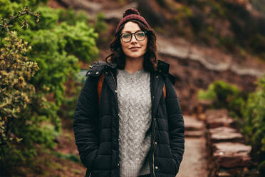 Porträt einer schönen jungen Frau in warmer Kleidung, die auf einem Bergpfad steht. Eine weibliche Touristin schaut in die Kamera, während sie auf den Stufen des Berges steht. - JLPSF16279