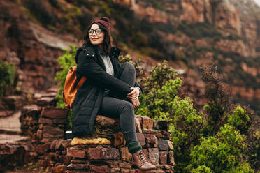 Weibliche Reisende sitzt auf Steinböschung am Berg und schaut weg. Frau in warmer Kleidung sitzt am Berghang. - JLPSF16280