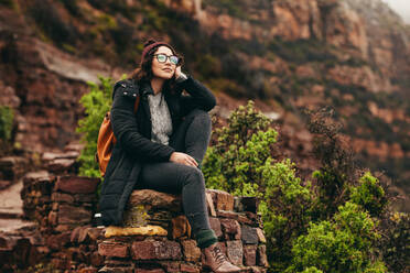 Entspannte Frau sitzt an der Seite eines Hügels und schaut auf die Landschaft. Weiblicher Reisender sitzt auf einem Stein mit Berg im Hintergrund. - JLPSF16281