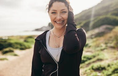 Lächelnde Frau in Sportkleidung, die nach dem Training im Freien steht. Fitness-Frau hört Musik mit Kopfhörern und schaut lachend in die Kamera. - JLPSF16293