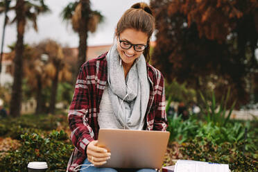Lächelnde junge Studentin, die auf einem Campus mit einem Laptop sitzt. Frau, die mit einem Kopfhörer Musik vom Laptop hört. - JLPSF16304