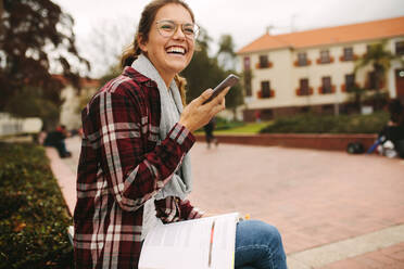 Lächelnde Studentin, die auf dem Universitätscampus mit einem Buch sitzt und mit einem Mobiltelefon spricht. Weibliche Studentin, die auf dem Campus telefoniert. - JLPSF16306