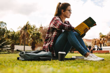 Seitenansicht einer jungen Frau, die im Freien auf einem College-Campus sitzt und ein Buch liest. Eine Studentin liest ein Buch auf einem Universitätscampus. - JLPSF16311