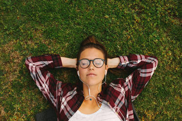 Draufsicht auf ein Mädchen mit Brille, das mit den Händen hinter dem Kopf und geschlossenen Augen im Gras liegt. Eine Studentin hört Musik mit Kopfhörern auf dem College-Campus. - JLPSF16313