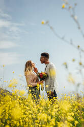 Seitenansicht eines jungen Mannes, der seine Freundin anschaut, die Blumen im Freien hält. Ein gemischtrassiges Paar steht zusammen und schaut sich liebevoll an. - JLPSF16318