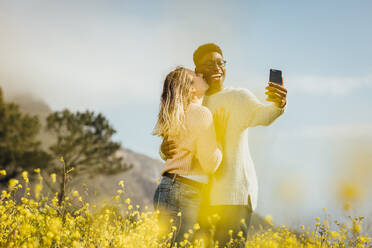 Romantisches Paar, das im Freien steht und ein Selfie macht. Ein afrikanischer Mann macht ein Selfie mit seinem Handy, während die Frau ihn auf die Wange küsst. - JLPSF16321