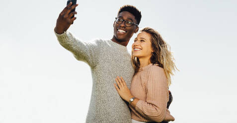 Verliebtes junges Paar im Sommerurlaub nimmt ein Selbstporträt mit Smartphone im Freien auf. Interracial Paar im Urlaub nimmt Selfie gegen hellen Himmel mit Handy. - JLPSF16322