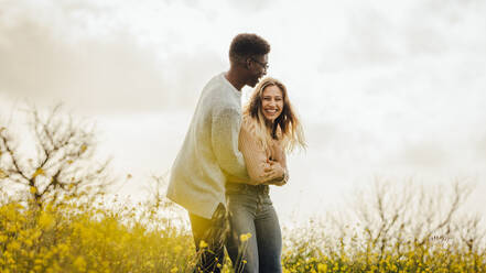 Ein Mann umarmt seine lächelnde Freundin von hinten. Ein Paar vergnügt sich auf einer blühenden Wiese bei Sonnenuntergang. - JLPSF16338
