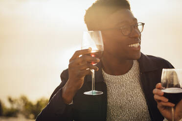 Junger afrikanischer Mann trinkt Wein, während er mit seiner Freundin im Freien sitzt. Paar genießt Wein bei einem Picknick. - JLPSF16345