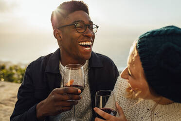 Smiling man and woman with wine glasses sitting in outdoors on a sunny day. Happy couple with red wine enjoying at picnic. - JLPSF16348