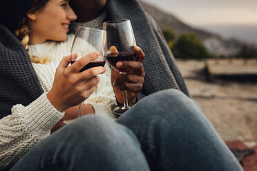 Cropped shot of a man and woman wrapped in blanket sitting together toasting wine while on a picnic. Couple enjoying a picnic outdoors with wine on a cold winter day. - JLPSF16352