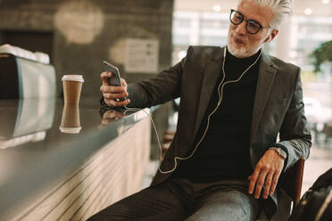 Älterer Geschäftsmann, der eine SMS auf seinem Smartphone schreibt, während er mit Kopfhörern in einem Café Musik hört. Ein Mann genießt die kostenlose drahtlose Internetverbindung in einem Café und sitzt vor dem Tresen. - JLPSF16362
