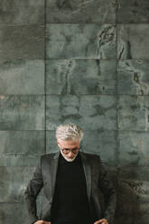 Vertikale Aufnahme eines gut aussehenden reifen Geschäftsmannes, der an einer grauen Wand steht und nach unten blickt. Ein älterer Mann in einer Jacke blickt nach unten. - JLPSF16375