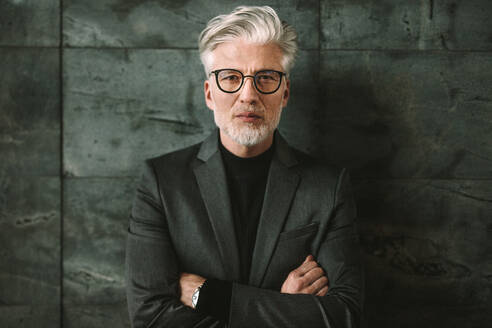 Portrait of confident mature businessman standing against a grey background. Handsome male in glasses standing with his arms crossed and looking at camera. - JLPSF16382