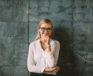 Portrait of beautiful young woman in formal wear standing against grey wall and smiling. Businesswoman with glasses looking at camera with hand on chin. - JLPSF16384