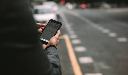 Nahaufnahme eines reifen Mannes, der eine Ride-Sharing-Anwendung auf seinem Smartphone nutzt, um ein Taxi auf einer städtischen Straße zu bestellen. - JLPSF16386